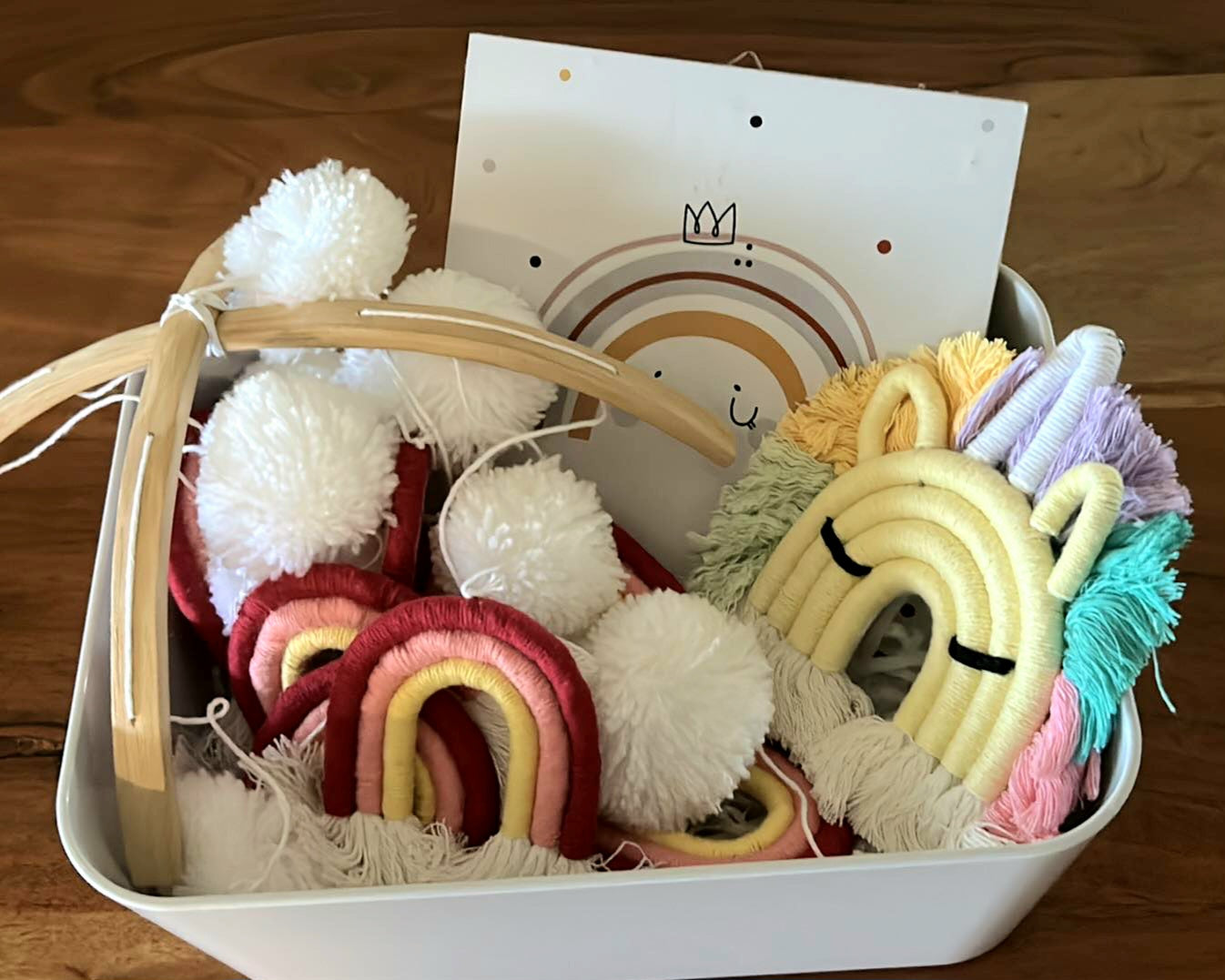 A basket containing a variety of colourful plush toys, including a unicorn keyring. a vibrant pom pom mobile, alongside a wall plaque with a rainbow crown design.