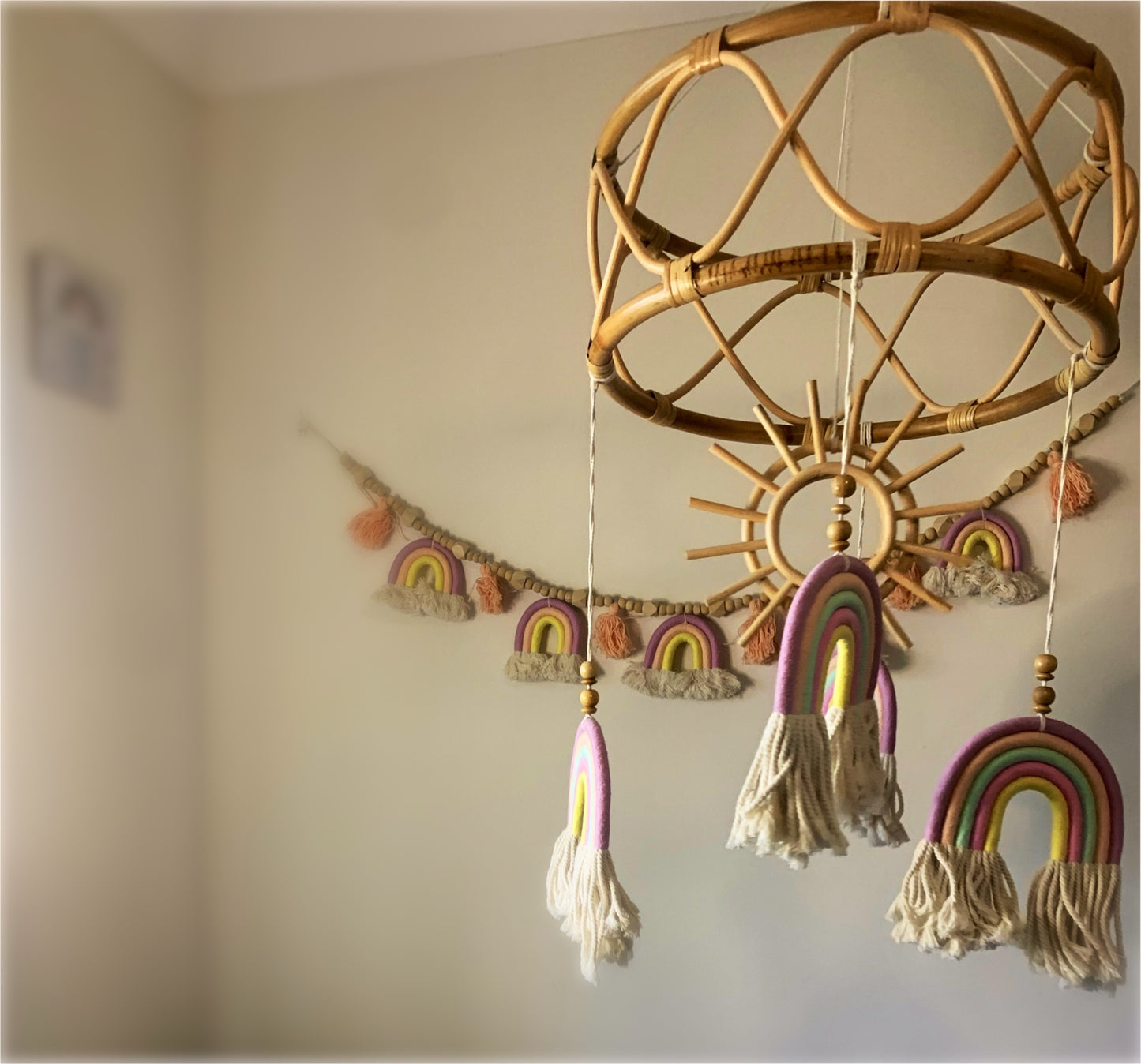 A handcrafted exclusive rattan mobile with a wooden sun and bamboo frame design, adorned with macrame rainbow decorations in a nursery setting.