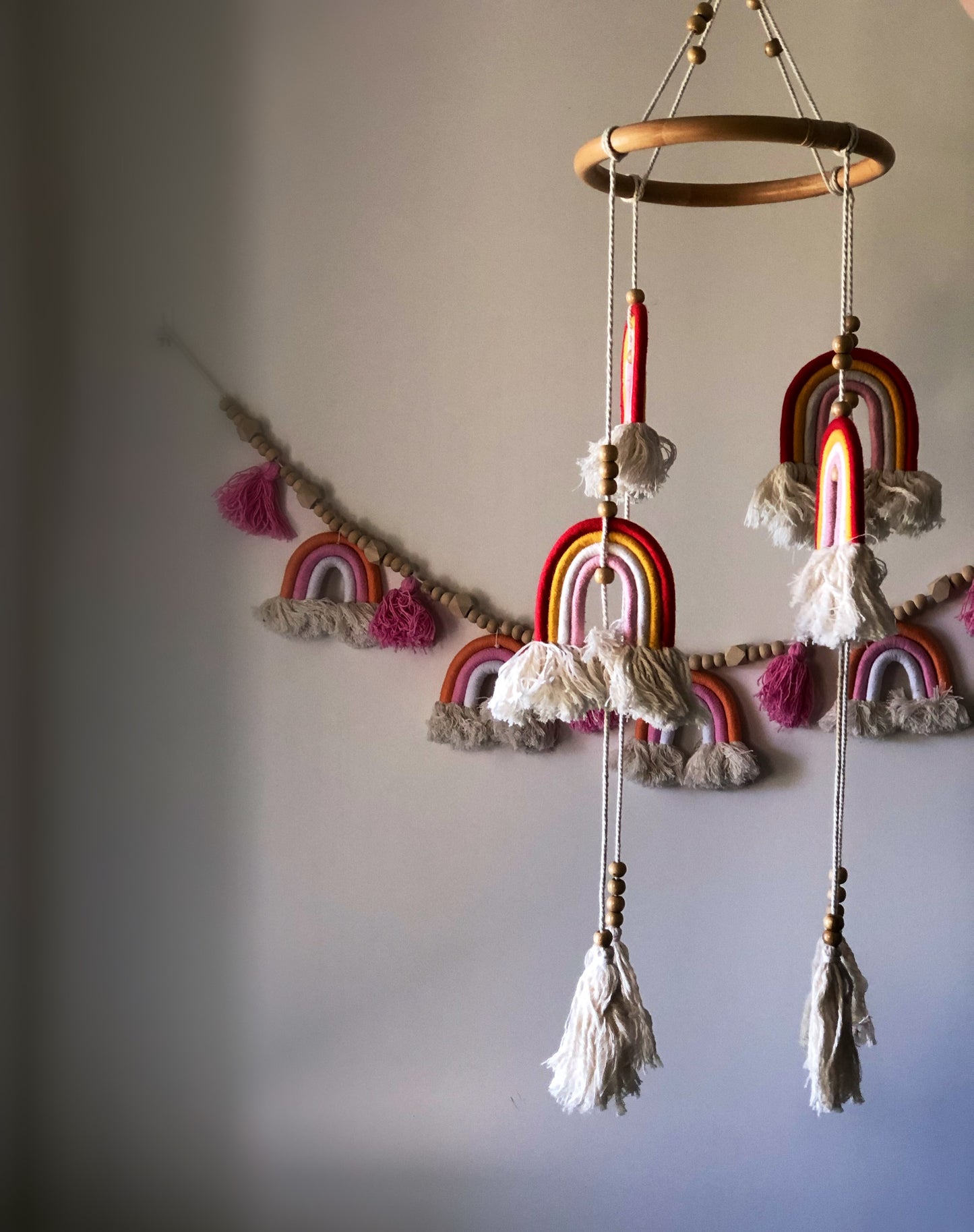 A long, hanging macrame rainbow mobile with multicolored stripes and tassels, displayed above a crib. Pink, yellow and red design rainbow