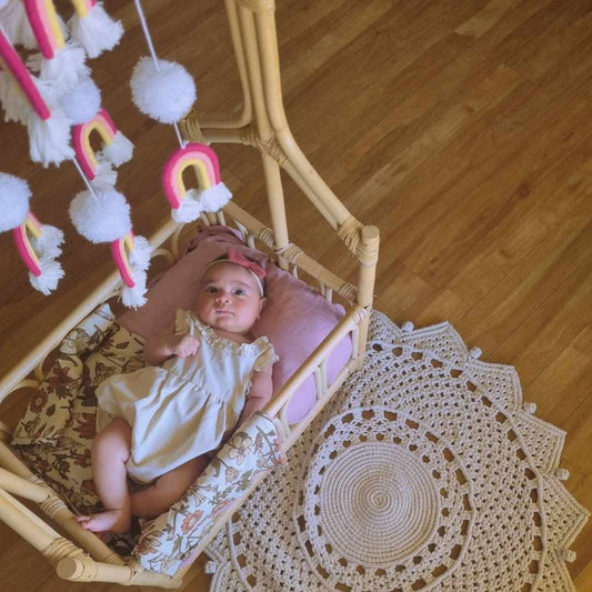 Macrame circular crochet mat beside a rattan bassinet with newborn baby and pom pom rainbow mobile — boho nursery floor rug Australia