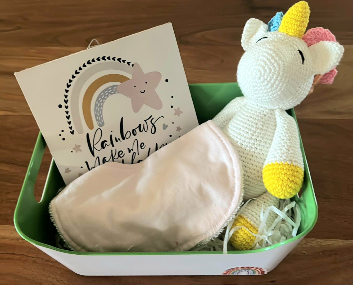 A basket containing a knitted white unicorn doll a pink bib, and a rainbow-themed plaque with the text 'Rainbows make me smile.'.