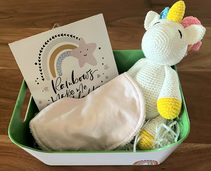 A basket containing a knitted white unicorn doll a pink bib, and a rainbow-themed plaque with the text 'Rainbows make me smile.'.