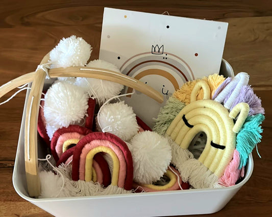 A basket containing a variety of colourful plush toys, including a unicorn  keyring. a vibrant pom pom mobile, alongside a wall plaque with a  rainbow crown design.