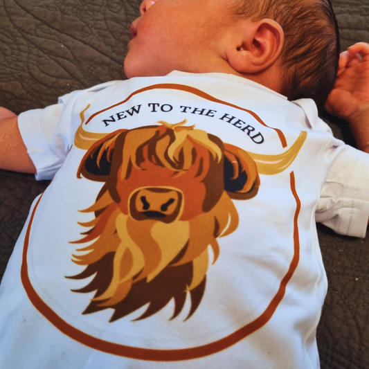 A baby wearing a white onesie with a highland cow design and the text 'NEW TO THE HERD' on the back.